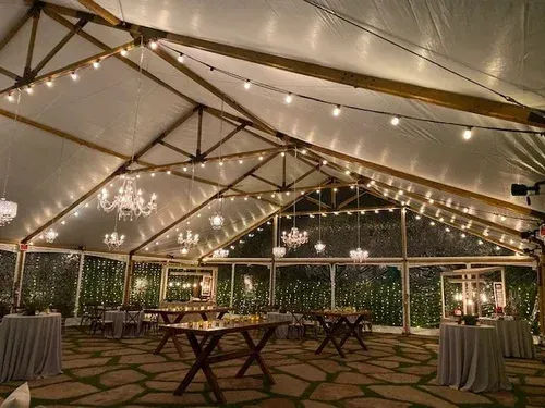Elegant reception tent with string lights, wooden tables, and round cocktail tables under a peaked canopy