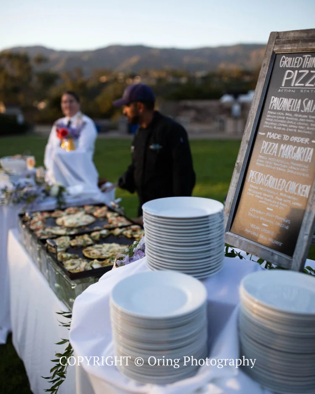 Outdoor pizza buffet with stacked plates and trays of pizza at a catering table