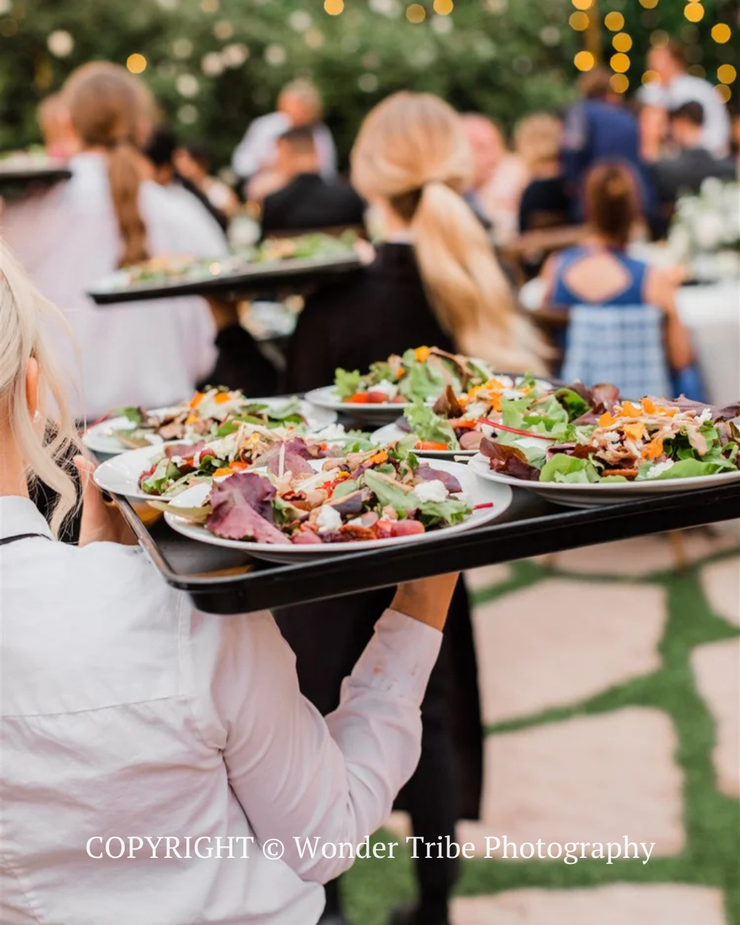 Server carrying trays of plated salad and food at an outdoor evening event with blurred guests and string lights