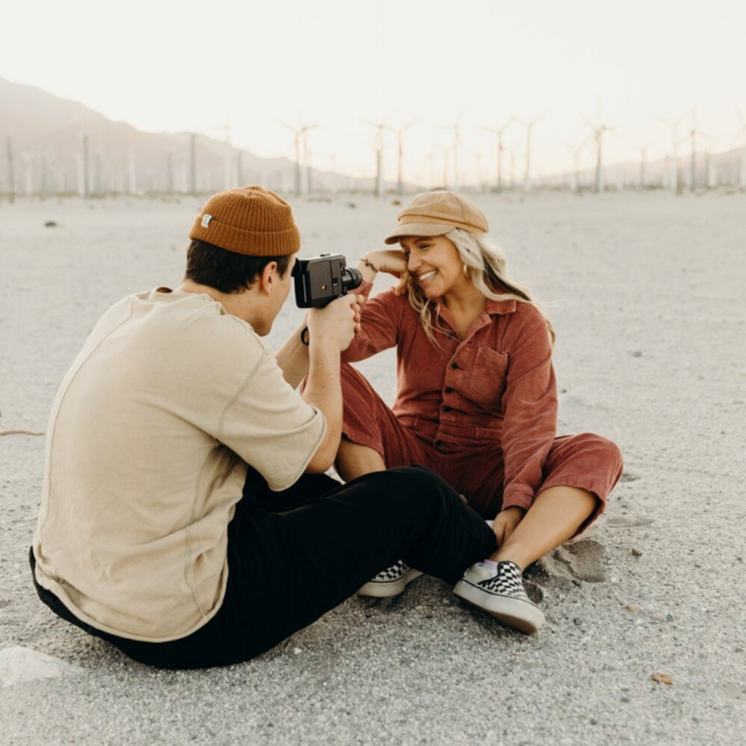 Two people sitting on gravel, smiling as one photographs the other at a beach with wind turbines in the distance