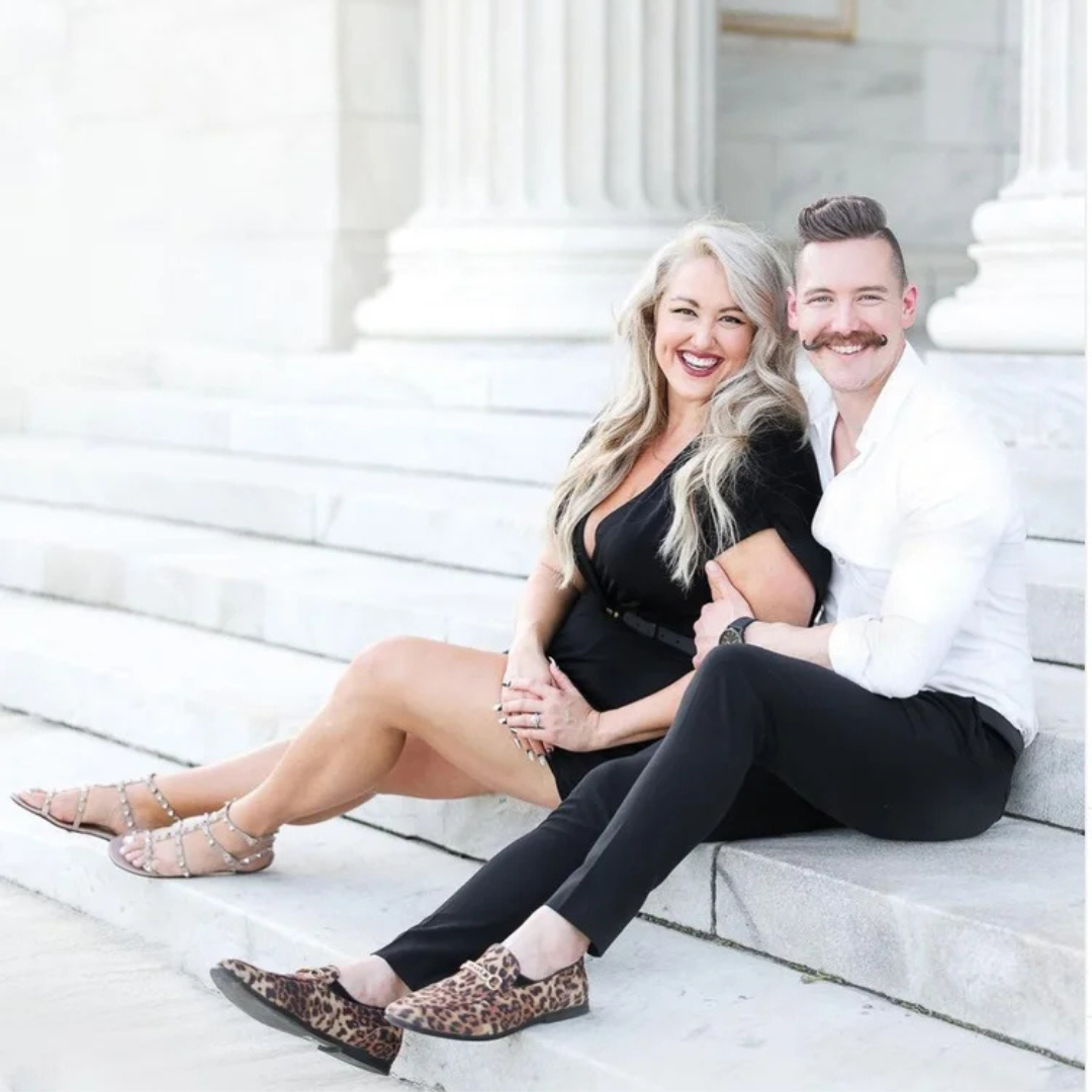 Smiling couple seated on white stone steps, dressed in black and white formalwear.