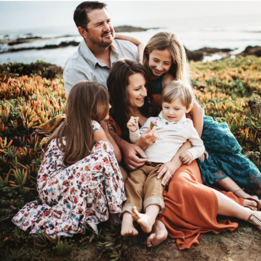 Family sitting together on grass by the ocean, smiling and embracing in a coastal field.