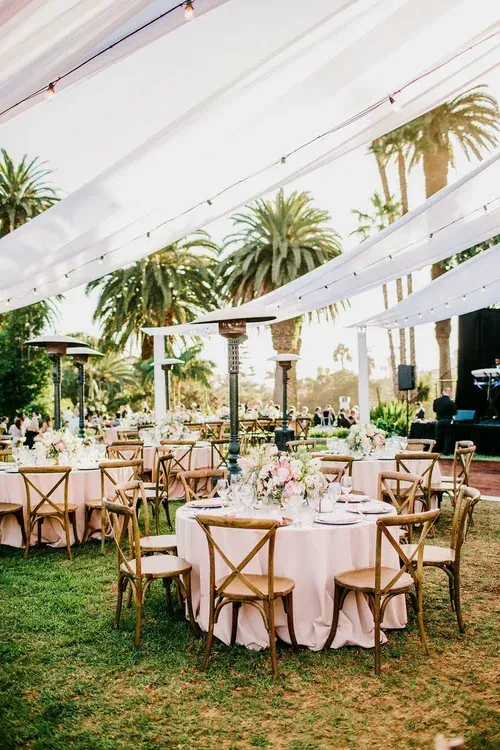 Outdoor wedding reception under white drapes with round tables, gold chairs, and greenery on a grassy lawn