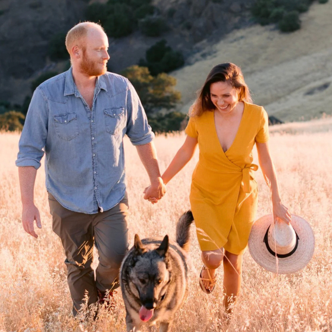 Couple walking hand in hand through sunlit grass with a dog, holding a straw hat