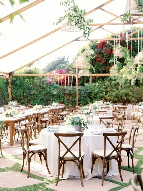 Elegant outdoor dining setup under a canopy with white tablecloths, wooden chairs, and lush floral greenery
