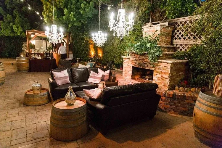 Cozy outdoor lounge with black sofas, barrel tables, hanging lights, and a brick fireplace at night