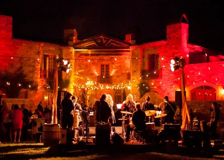 Outdoor evening party at a red-lit stone courtyard with people mingling at tables and a lit building backdrop
