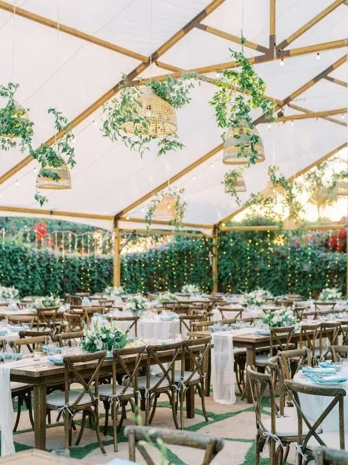 Decorated outdoor dining tent with wooden tables, white chairs, and hanging greenery under a peaked canopy