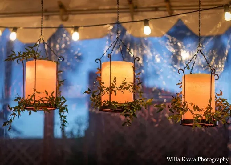 Three glowing lanterns hang under a canopy at dusk, with blurred trees and lights in the background.