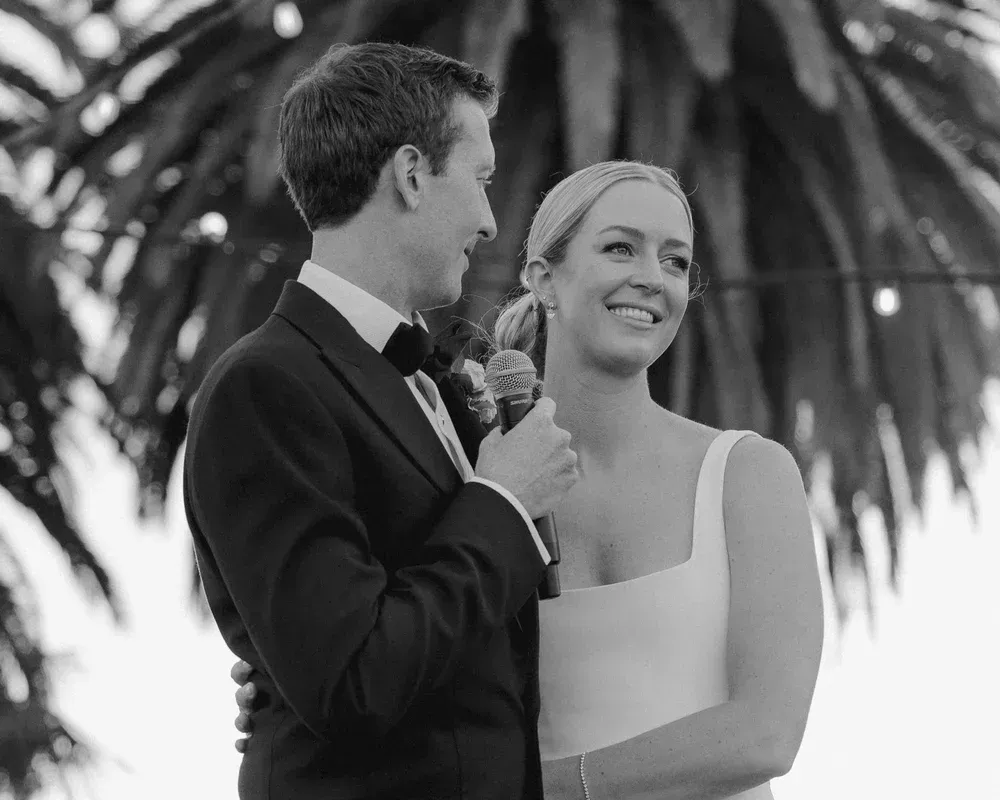 Couple in formal attire smiling during an outdoor wedding ceremony, with palm trees in the background