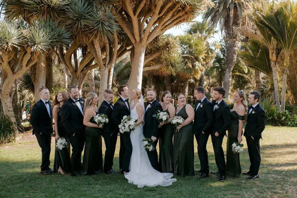 Wedding party posing outdoors under palm trees, with bride in white and attendants in black attire.