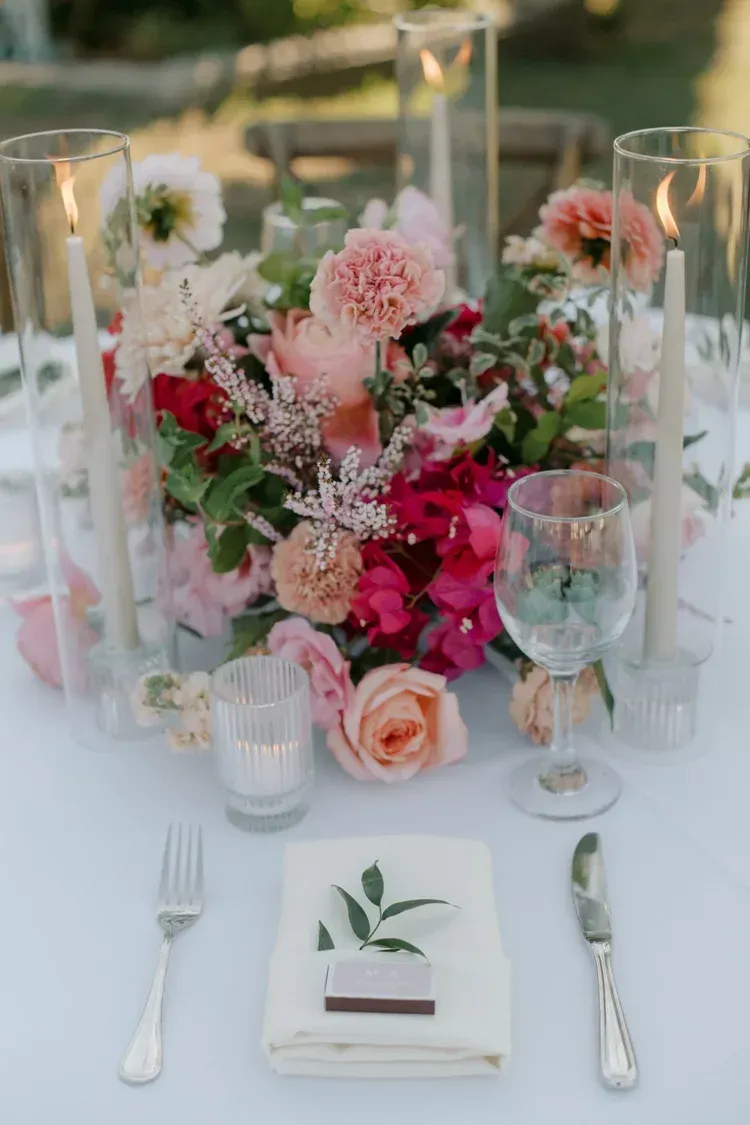 Elegant outdoor table setting with pink floral centerpiece, candles, glassware, and a folded napkin with greenery and silverware