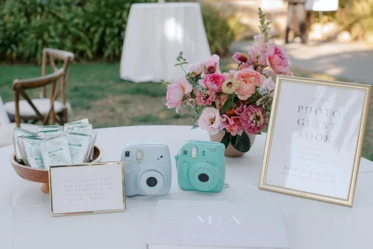 Wedding welcome table with pastel instant cameras, floral bouquet, and framed sign outdoors