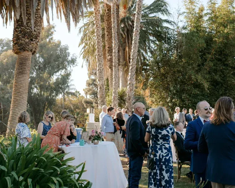 Guests mingle outdoors at a palm-lined evening event with cocktail tables and white linens.