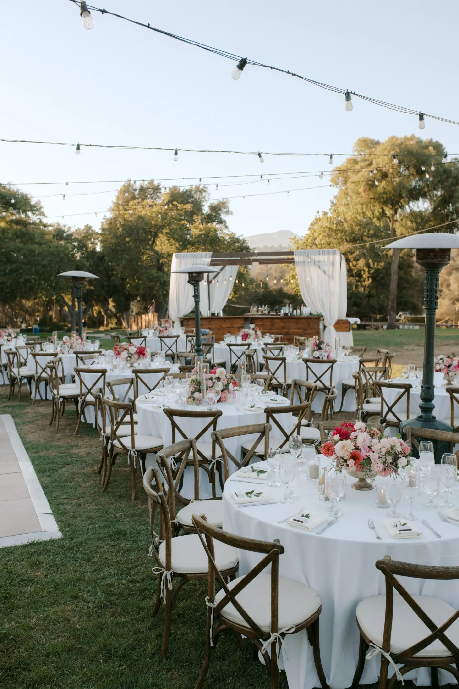 Outdoor wedding reception with round tables, white chairs, floral centerpieces, and string lights on a lawn