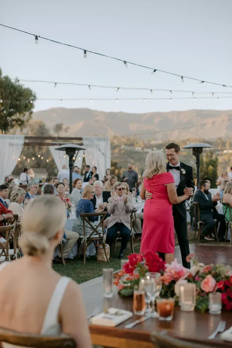 Outdoor wedding reception with a couple dancing among seated guests under string lights at sunset