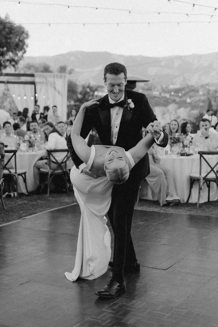 Couple dancing outdoors at a formal reception, with guests seated at tables in the background.