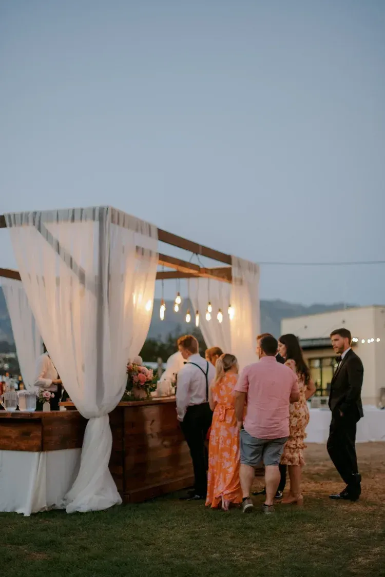 Guests gathered around a lit outdoor bar under draped white fabric at dusk.
