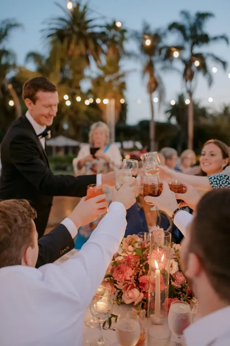 Guests raise glasses at an outdoor evening wedding reception with palm trees and string lights.