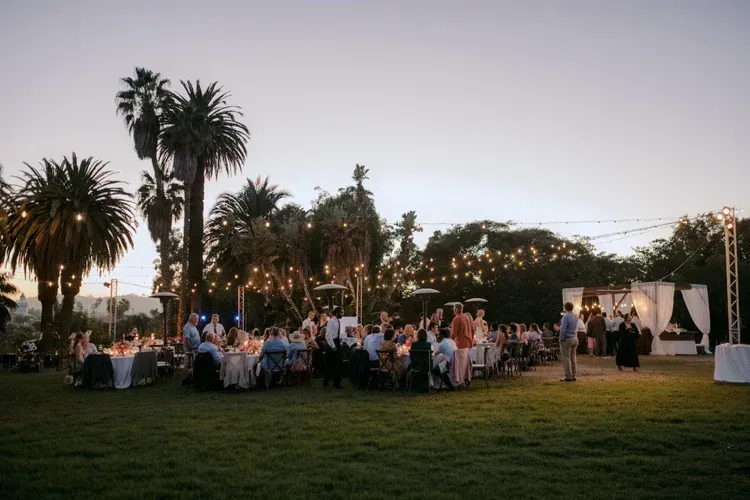 Outdoor evening reception with guests seated at round tables on a grassy lawn beside palm trees and string lights