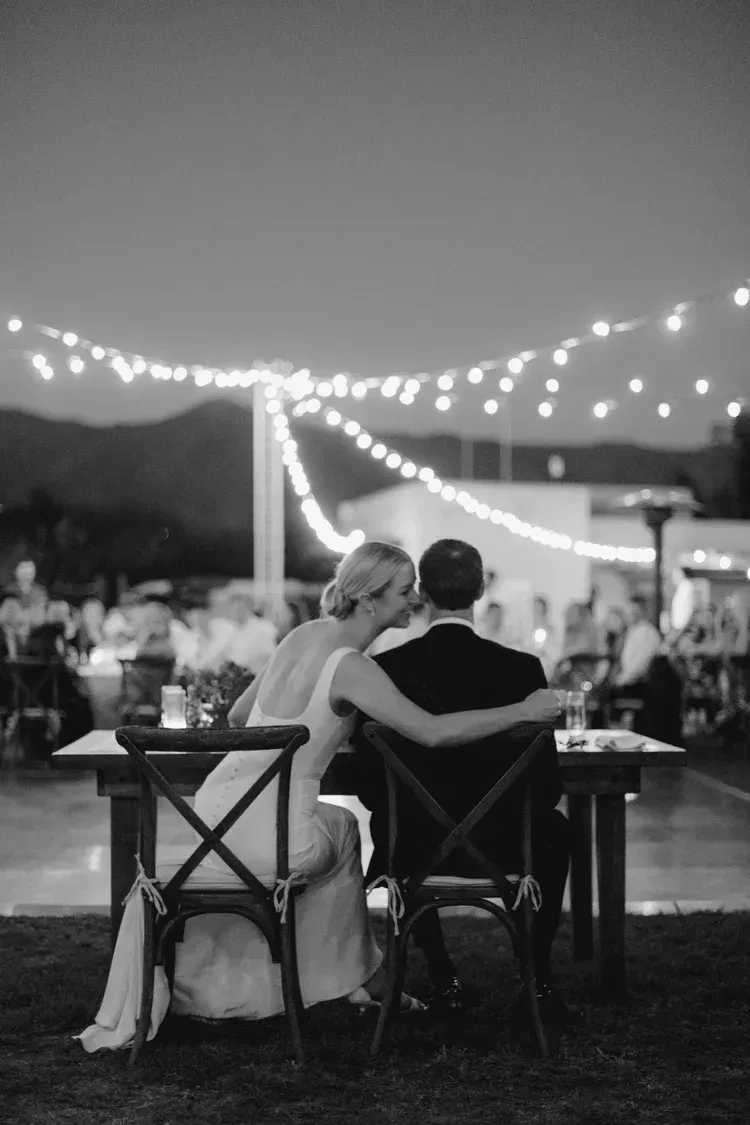 Couple seated at a candlelit outdoor dinner, embracing under string lights at dusk