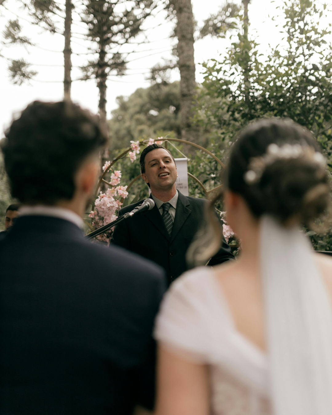 Outdoor wedding ceremony with officiant speaking to couple, bride seen from behind, trees in background