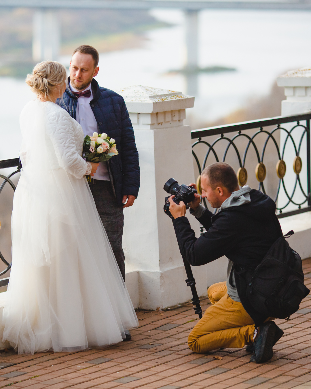 Wedding photographer kneels to photograph a bride and groom on a waterfront terrace.