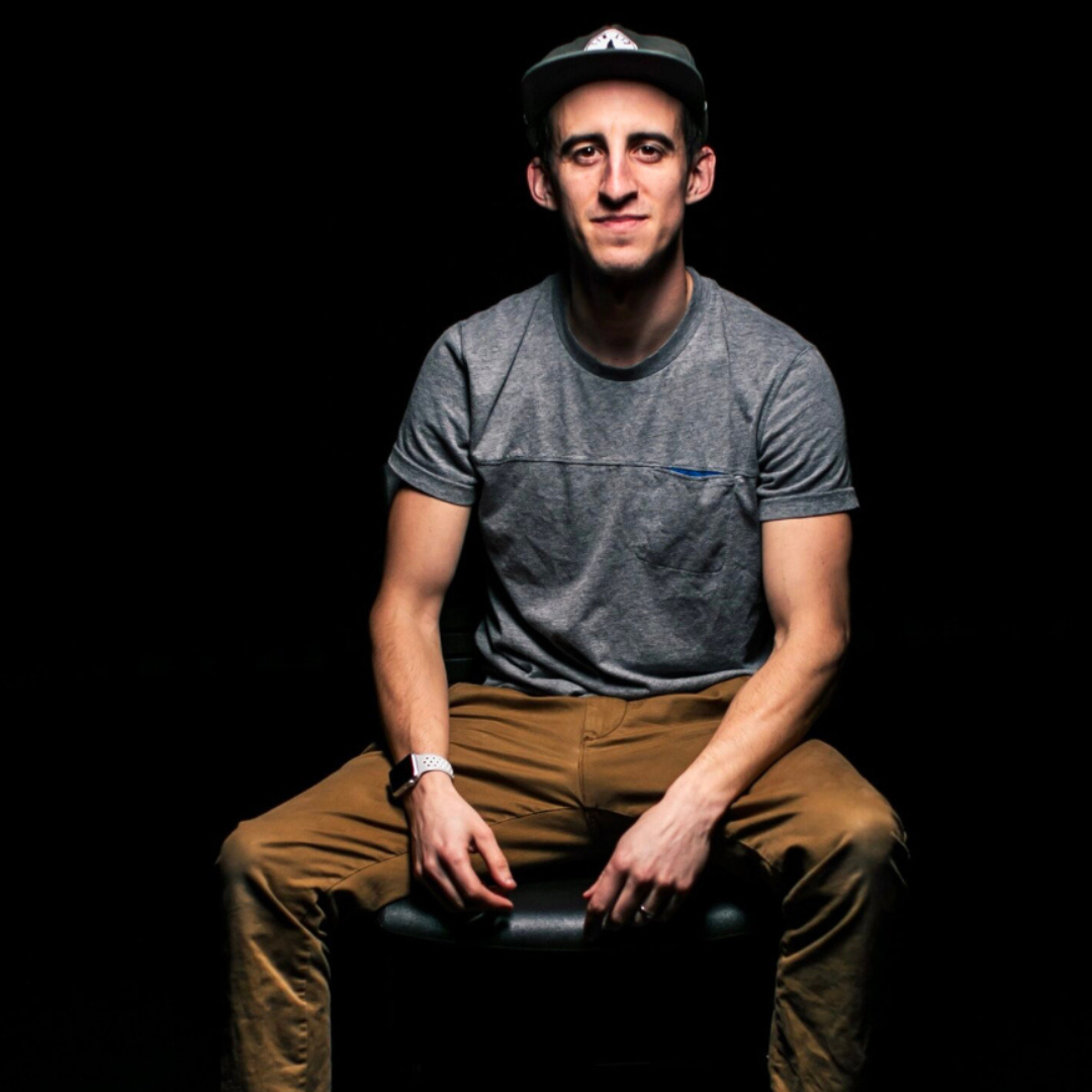 Man seated on a stool against a black background, wearing a gray T-shirt, tan pants, and a cap.
