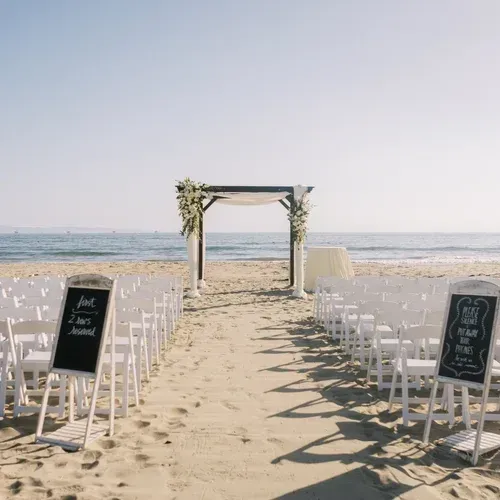 Beach wedding aisle with white chairs, floral arch, and chalkboard signs facing the ocean.