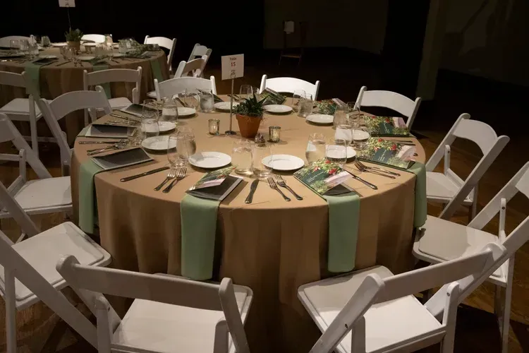 Round banquet table set with white folding chairs, tan and green tablecloth, and dishes for a formal event