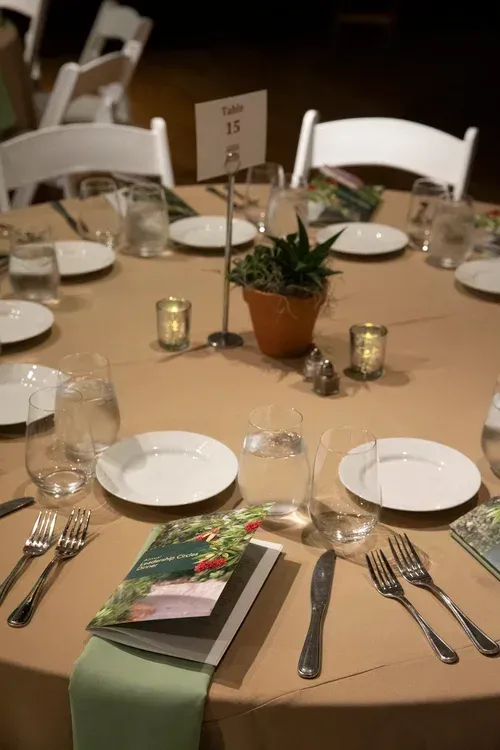 Round banquet table set with white plates, wine glasses, candles, and a small potted plant centerpiece.