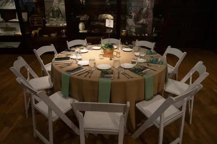 Round banquet table set with white chairs, green and tan tablecloth, and place settings in a dimly lit room