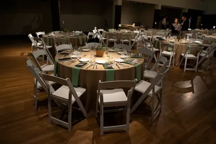 Round banquet tables and white folding chairs arranged in a dimly lit event hall.