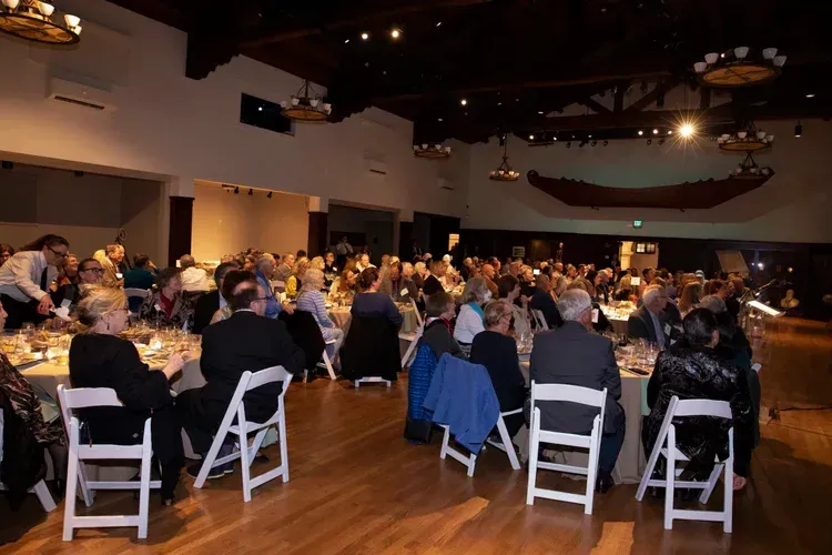 Guests seated at round tables in a warmly lit banquet hall, watching a presentation.