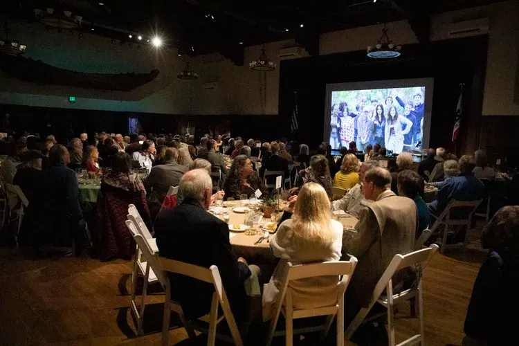 Audience seated at round tables watching a presentation on a large screen in a dim conference hall