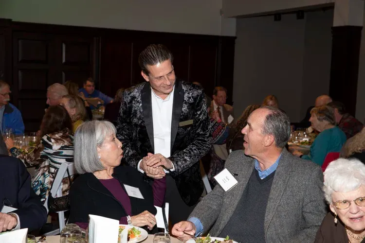 Man speaking with seated guests at a formal indoor event, with dark wood paneling and table settings