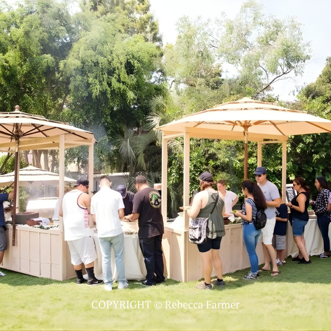 People lined up at outdoor beige drink stations under tents in a sunny garden.