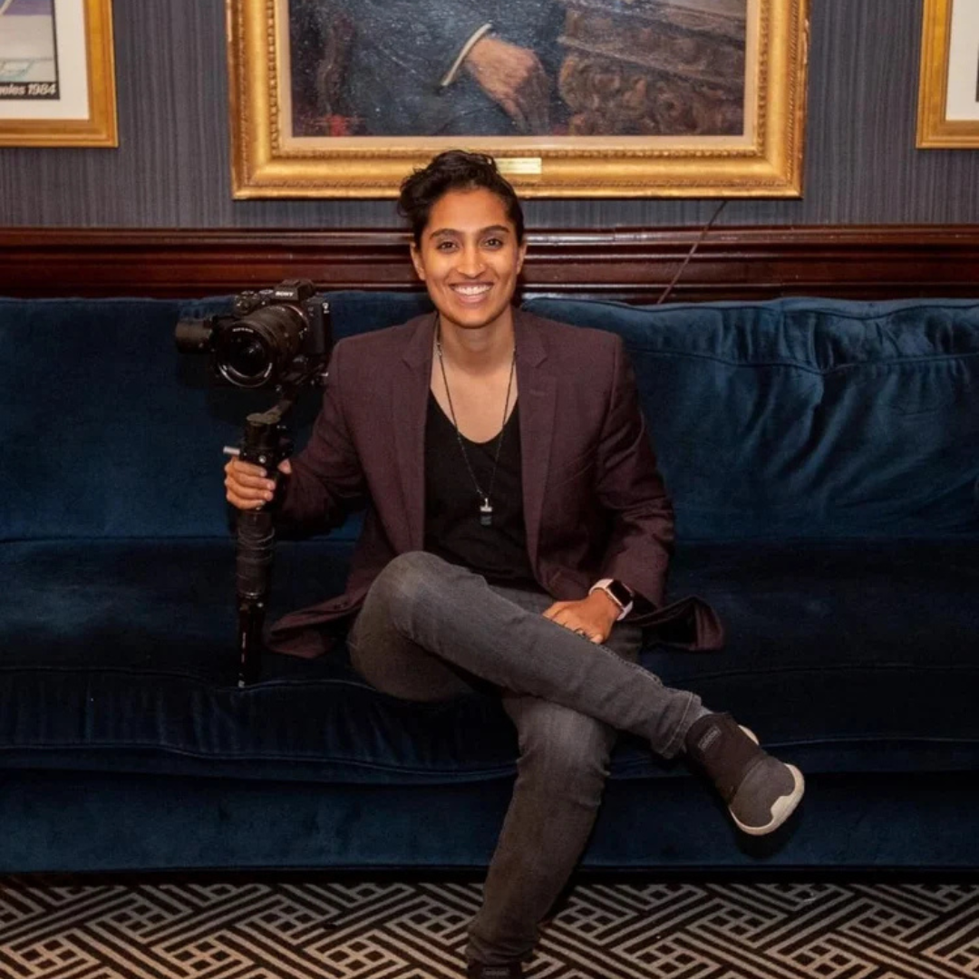 Person sitting cross-legged on a blue velvet couch, smiling and holding a camera rig.