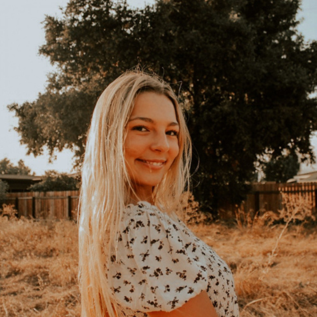 Smiling blonde person in a white floral top standing outdoors near a large tree at sunset