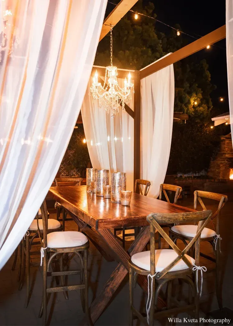 Outdoor dining table under draped white curtains and a chandelier, set for a romantic evening meal.