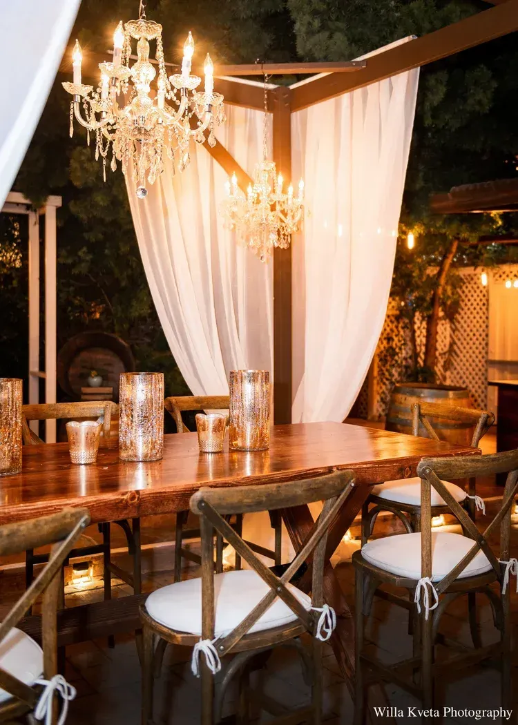 Elegant outdoor dining setup with chandelier lighting, white drapes, and a wooden table and chairs at night