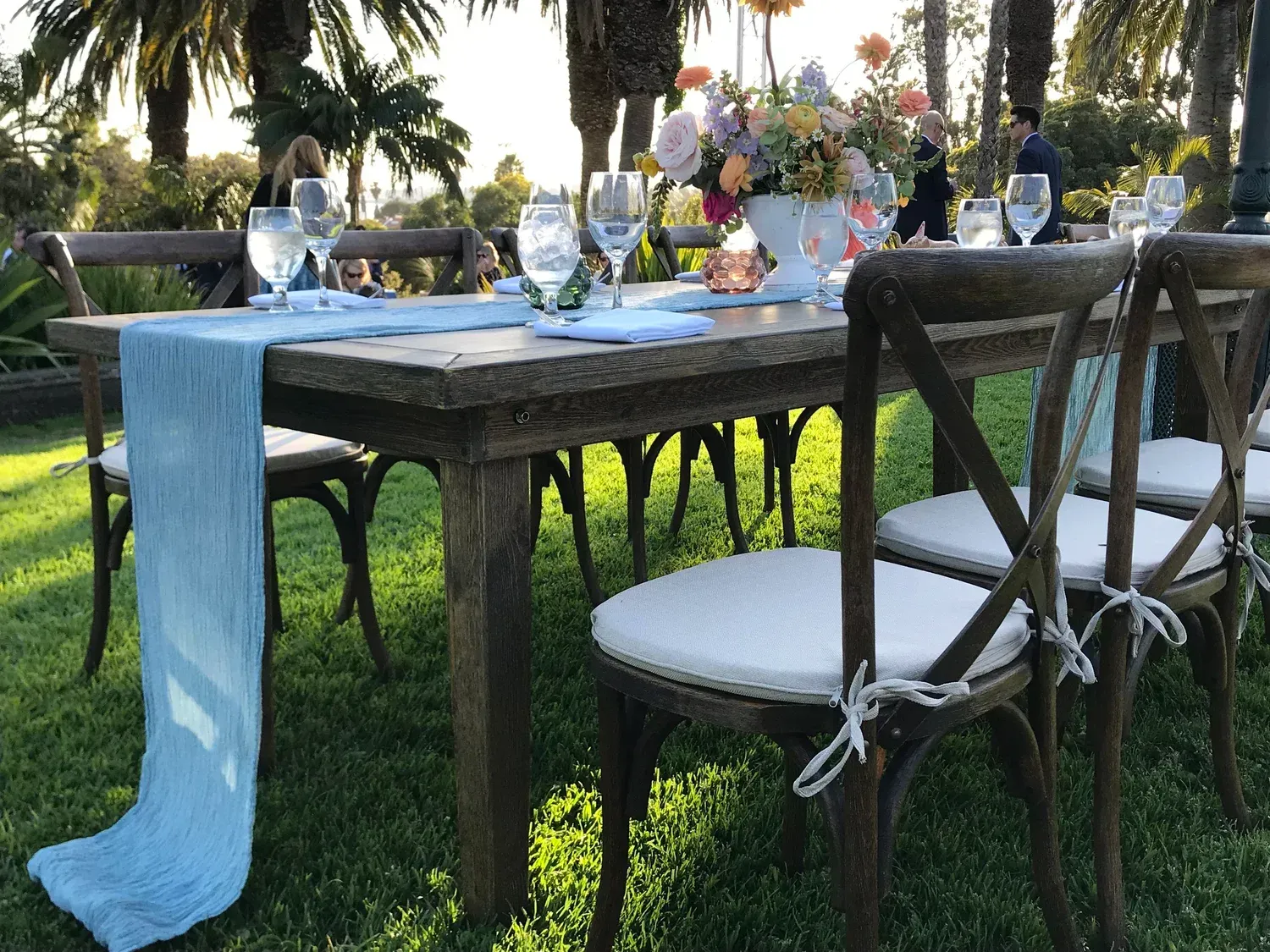 Outdoor dining table set on grass with white chairs, blue table runner, and floral centerpiece under palm trees