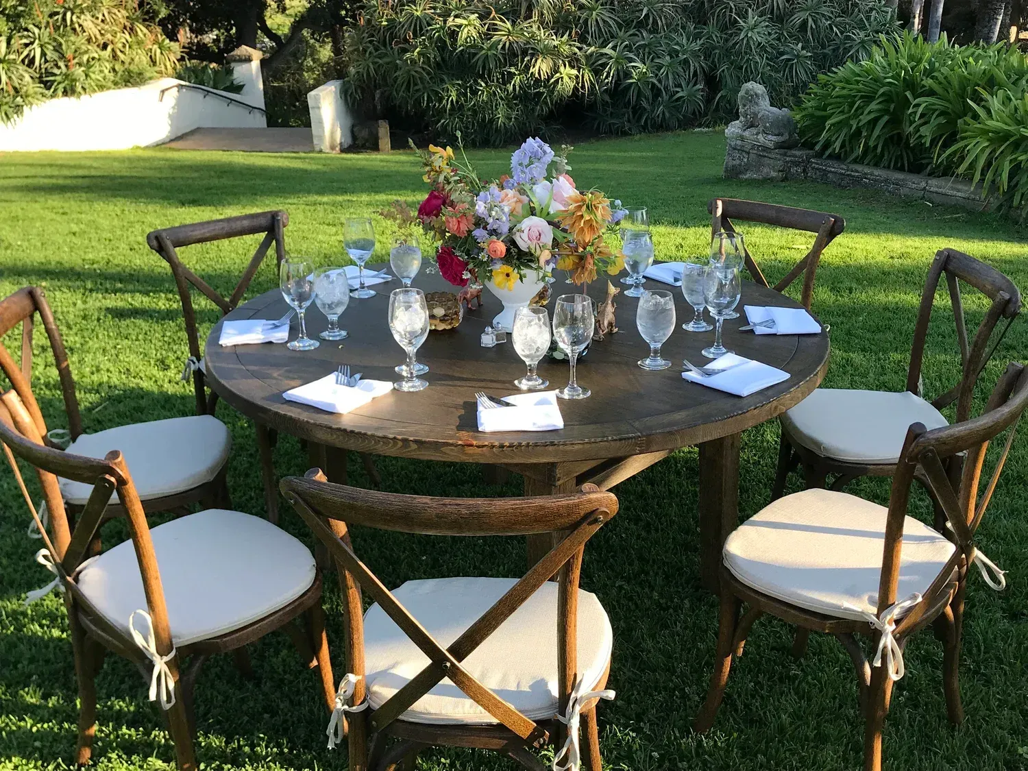 Outdoor round table set with white-cushioned wooden chairs, floral centerpiece, and glassware on a grassy lawn