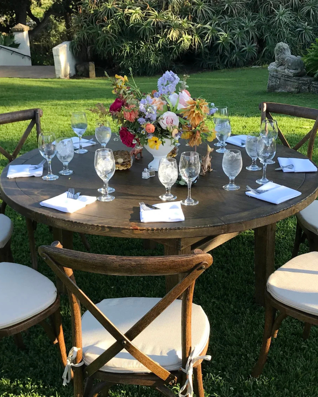 Round outdoor dining table set with glasses, napkins, and a colorful flower centerpiece in a garden