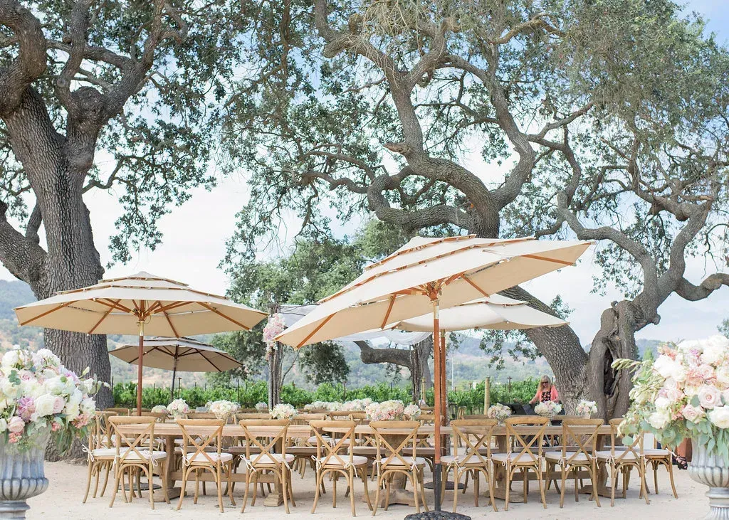 Outdoor cafe patio with tan umbrellas, wooden tables, and large trees in the background