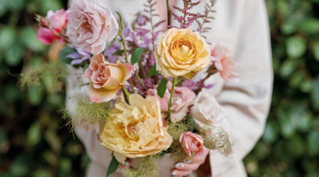 Person holding a bouquet of pastel roses and greenery, with a soft white sweater in the background