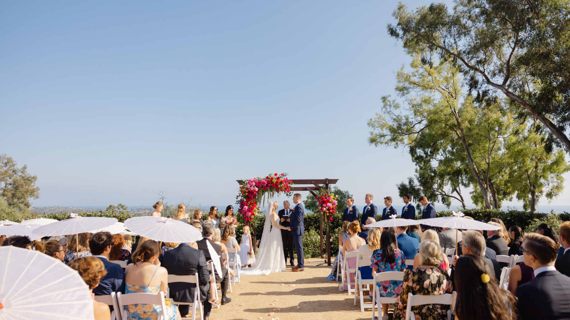 Outdoor wedding ceremony with bride and groom under a red floral arch, guests seated under white umbrellas