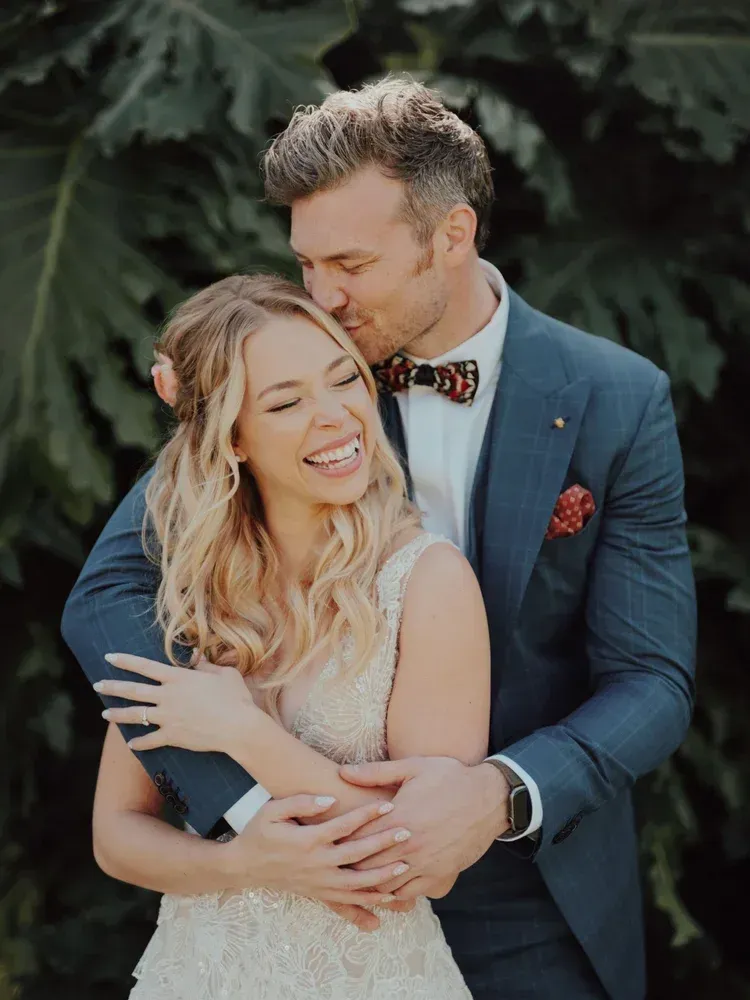 Couple embracing outdoors, smiling and wearing formal attire against a leafy background