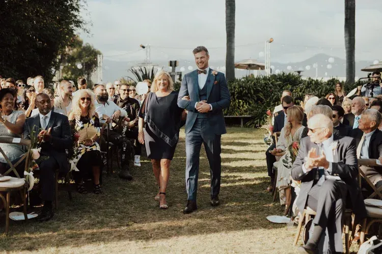 Couple walking down an outdoor wedding aisle between seated guests on a sunny day