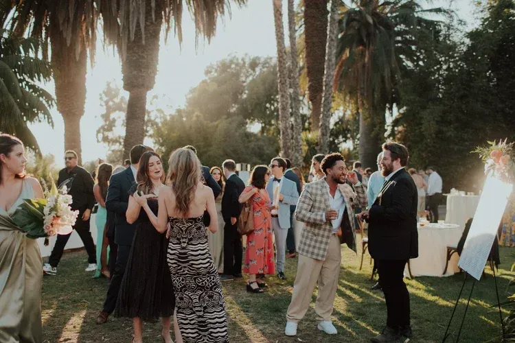 Guests mingle at an outdoor evening event among palm trees, with a man speaking near a bright light panel.